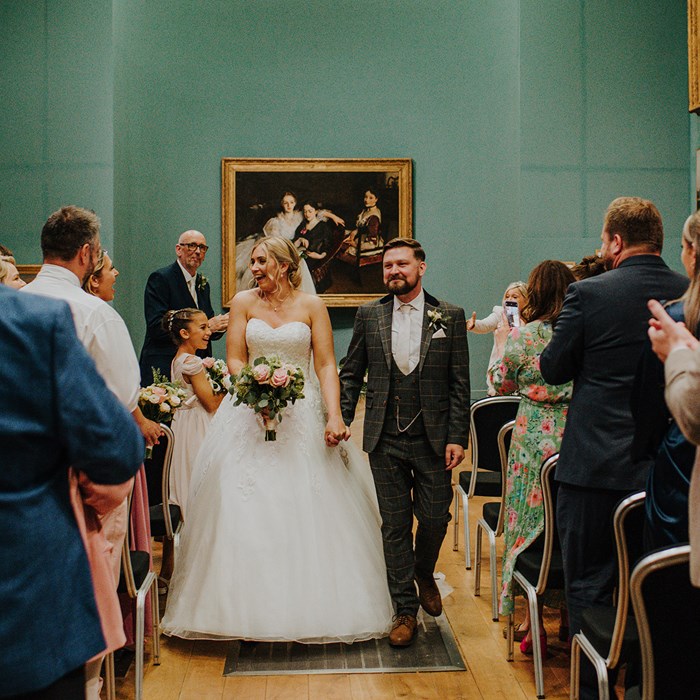 A bride in a wedding dress and groom in a suit walk down an aisle between rows of standing, clapping guests. In teh background is a historic painting of three women.