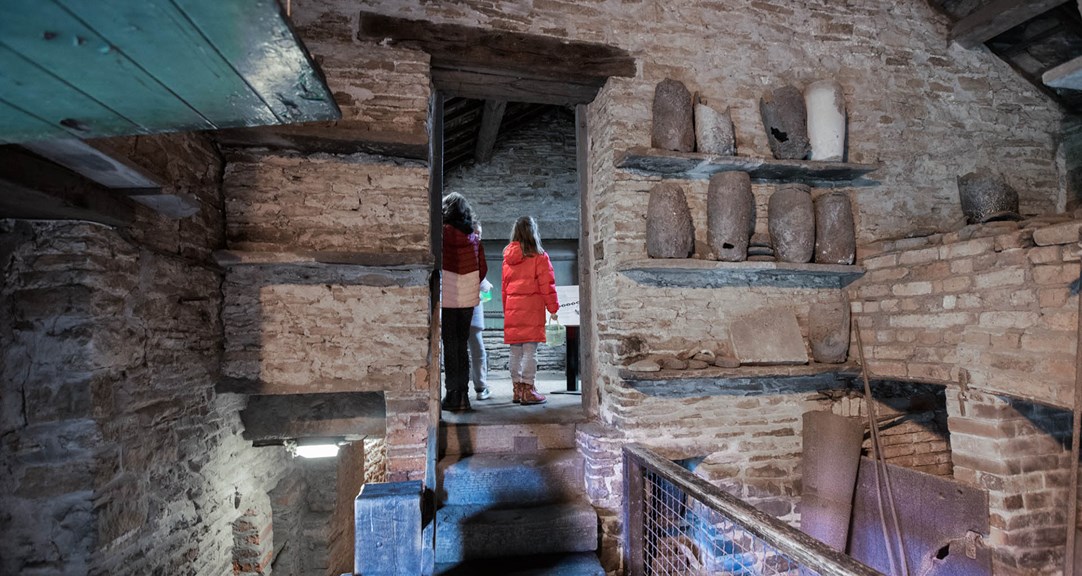 Visitors seen through a doorway with a display of old crucibles on very old wall shelves off to one side.