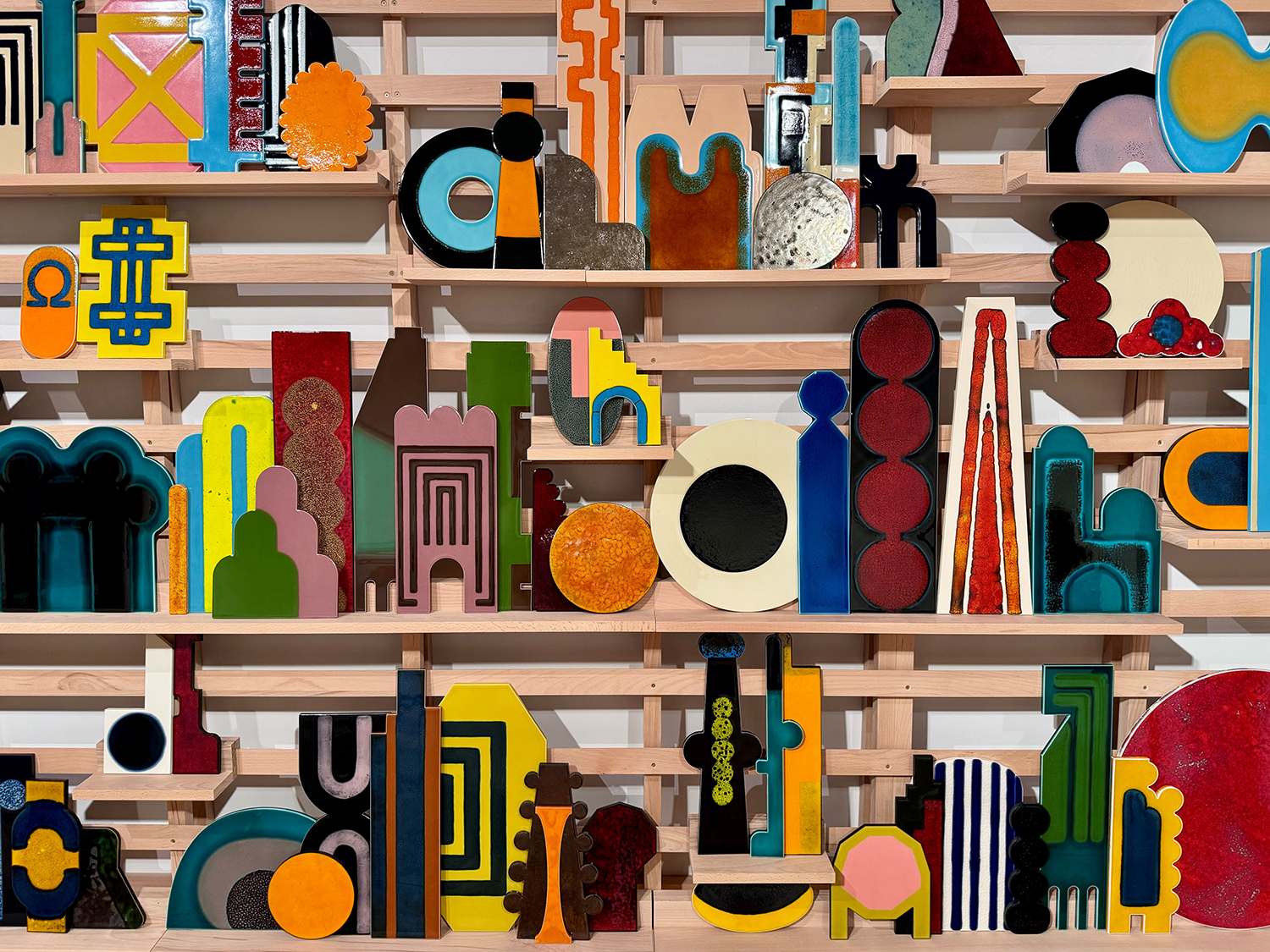 A series of glazed ceramic geometric shapes in bold, bright colours arranged on a wooden shelving structure.