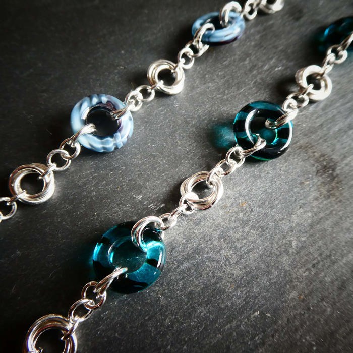 A close-up photograph of two silver chains. Some of the links are silver chain, some are thicker, blue, and made from a slightly transparent material. They are photographed against a grey background.