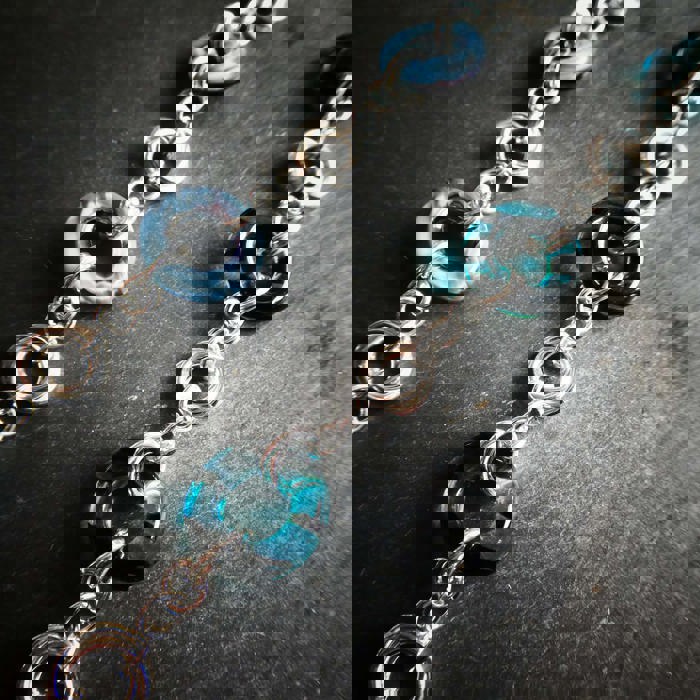 A close-up photograph of two silver chains. Some of the links are silver chain, some are thicker, blue, and made from a slightly transparent material. They are photographed against a grey background.