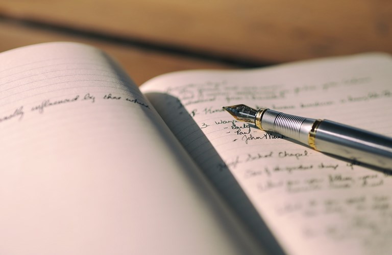 A fountain pen laid on an open, lined notebook which has indecipherable handwriting on the page. 