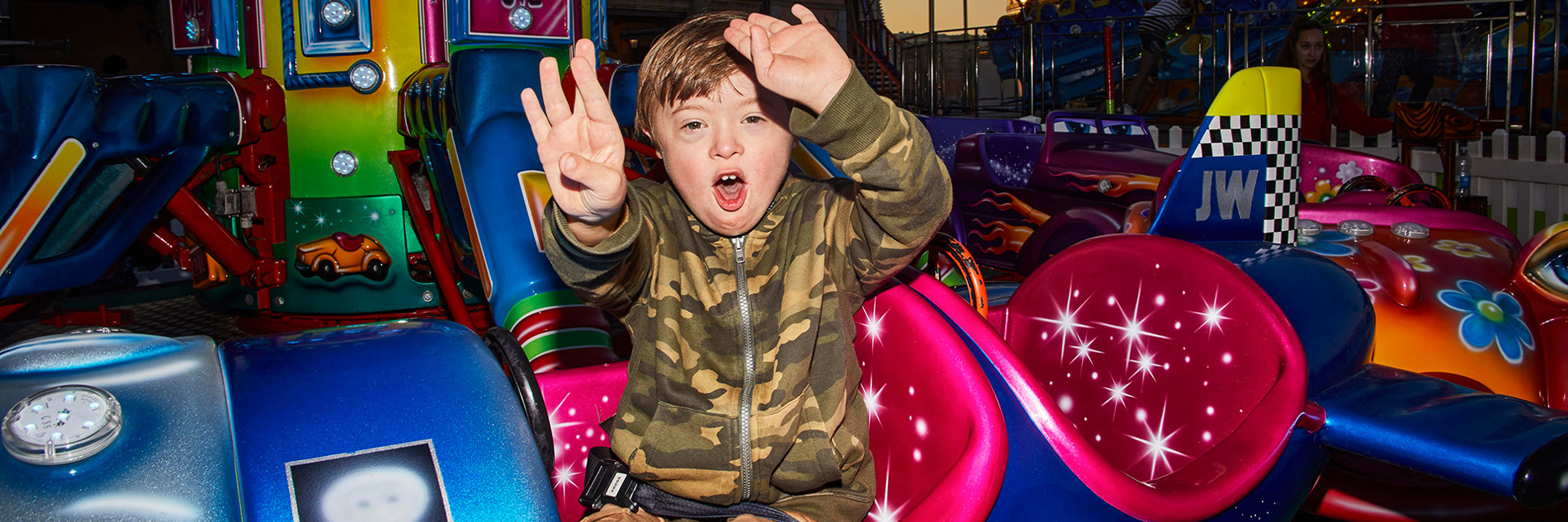 A photograph of a young boy sat on a fairground ride. He is sat in an airplane with a happy expression on his face, raising both arms up.