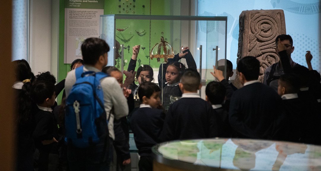 A large group of primary school students in the Beneath Your Feet gallery, gathered around a display case containing anglo-saxon artefacts. 