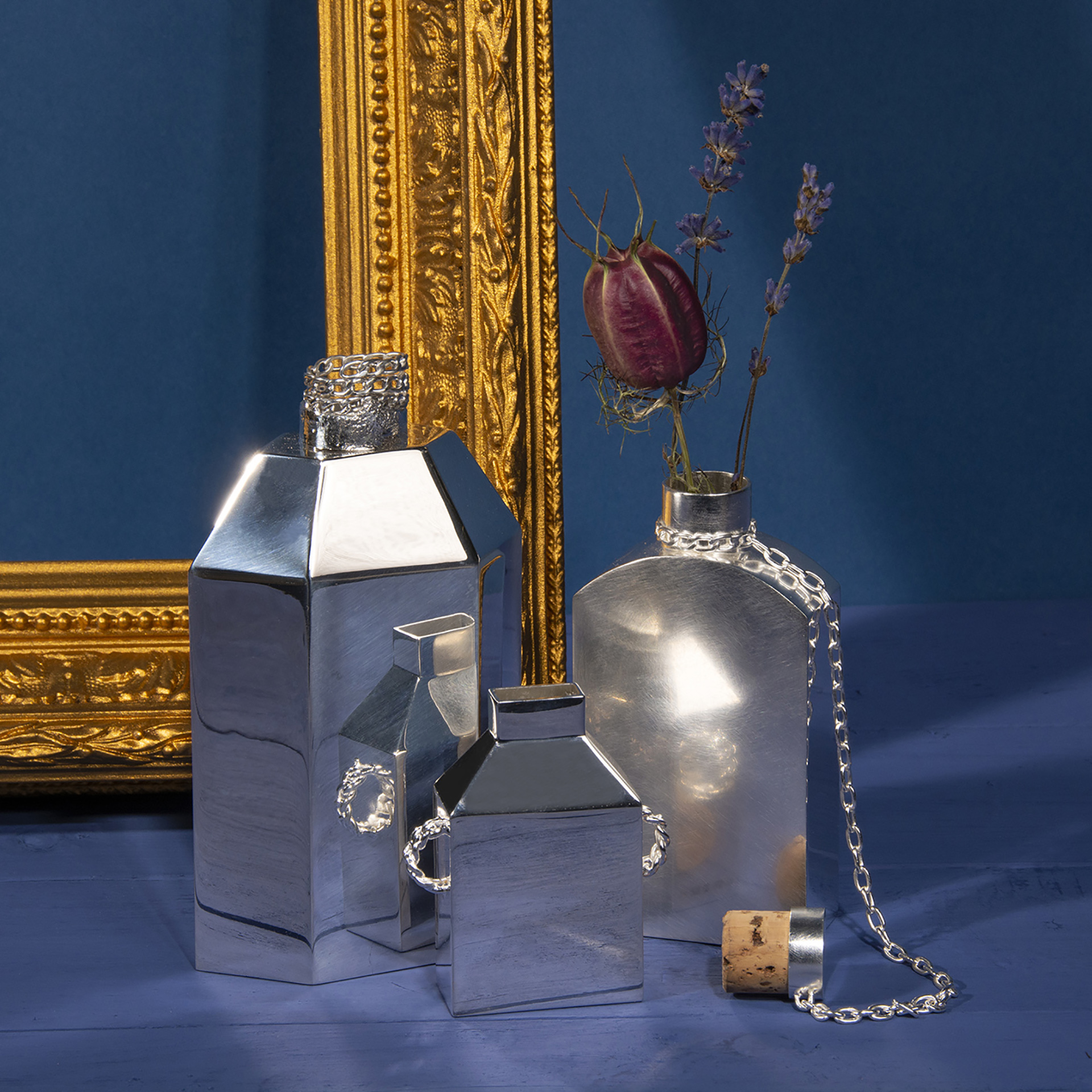 Three metal vases sitting next to a golden frame on a blue background. One of the vases is holding purple flowers. 