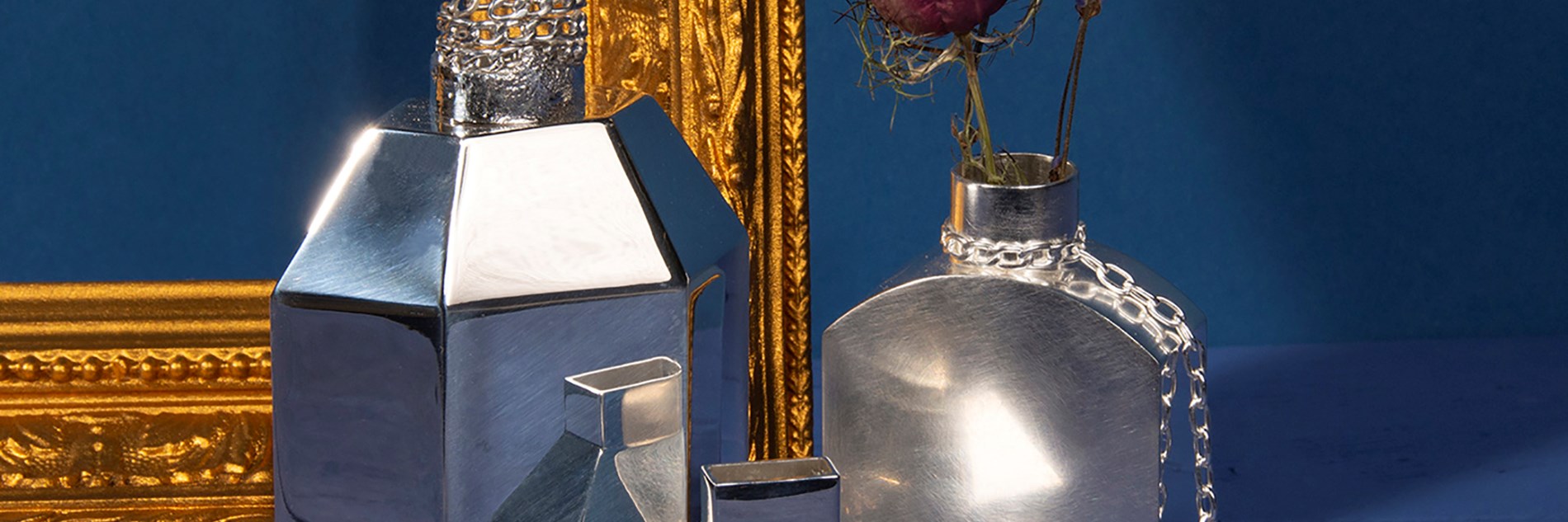 Three metal vases sitting next to a golden frame on a blue background. One of the vases is holding purple flowers. 