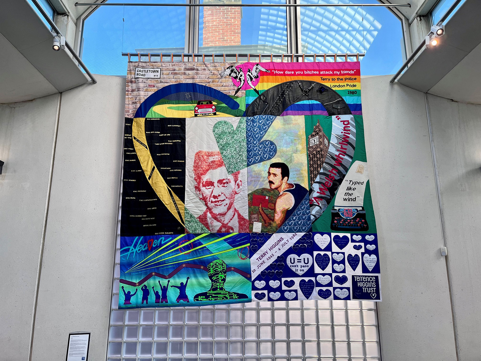 A colourful embroidered quilt featuring text and images. At the centre are two portraits of Terry Higgins which are framed by a heart motif. The quilt is hung on a concrete and glass wall in a contemporary building.
