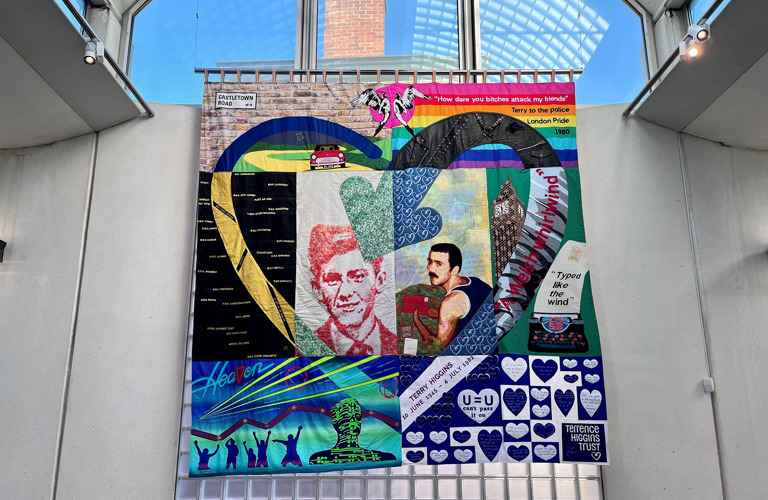 A colourful embroidered quilt featuring text and images. At the centre are two portraits of Terry Higgins which are framed by a heart motif. The quilt is hung on a concrete and glass wall in a contemporary building.