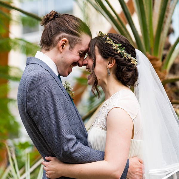 A smiling bride in a wedding dress and a groom in a suit face each other in front large exotic plants.