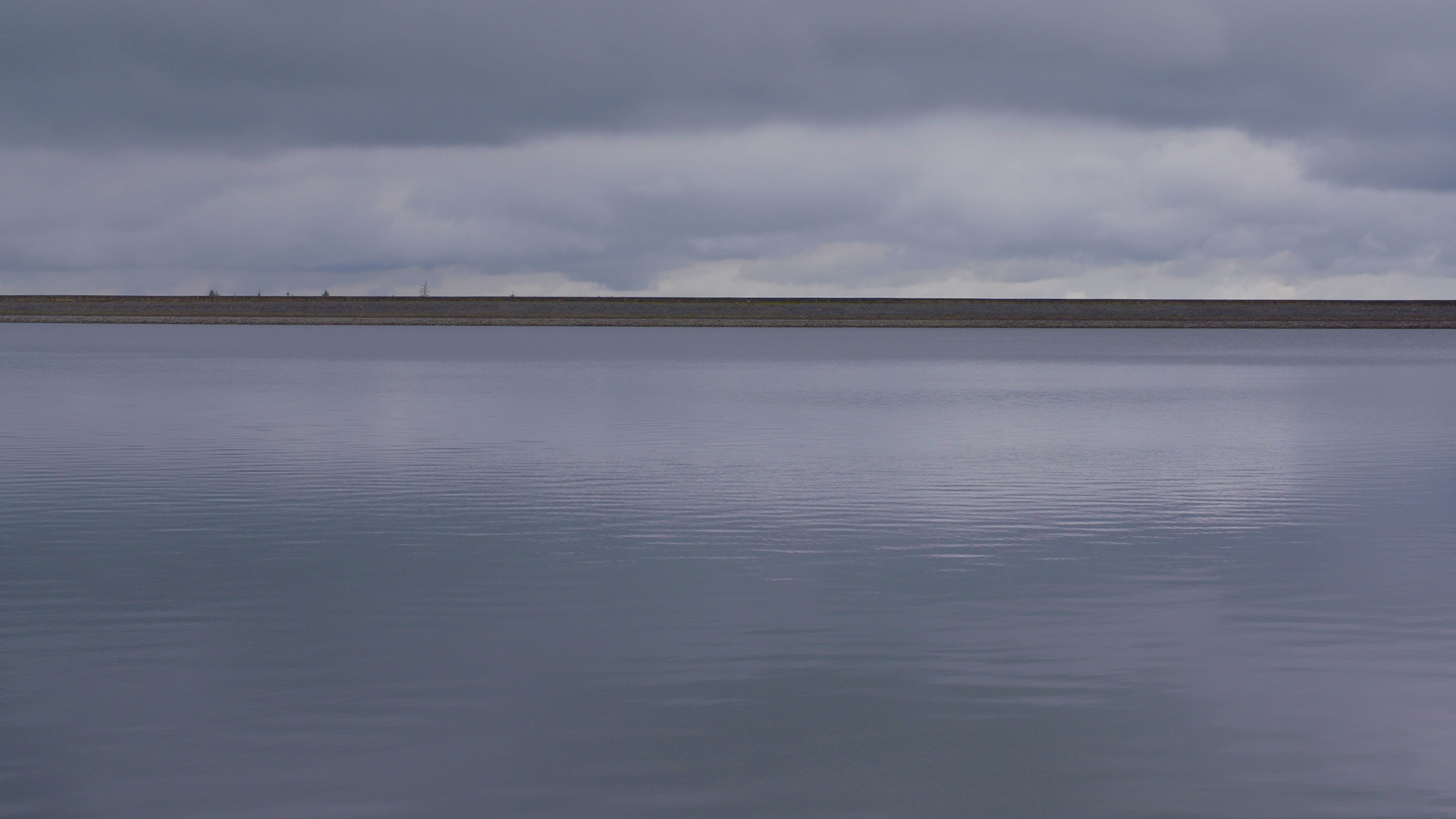A photograph of a reservoir meeting the horizon. The sky is overcast.
