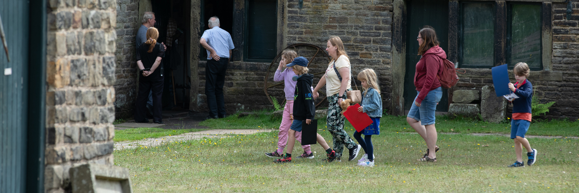 A group of adults and children walk across a grassed area in front of a row of single story historic stone buildings. 