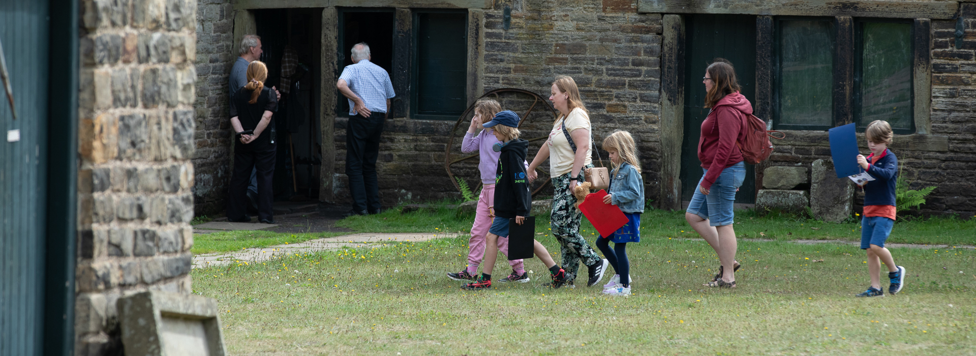 A group of adults and children walk across a grassed area in front of a row of single story historic stone buildings. 