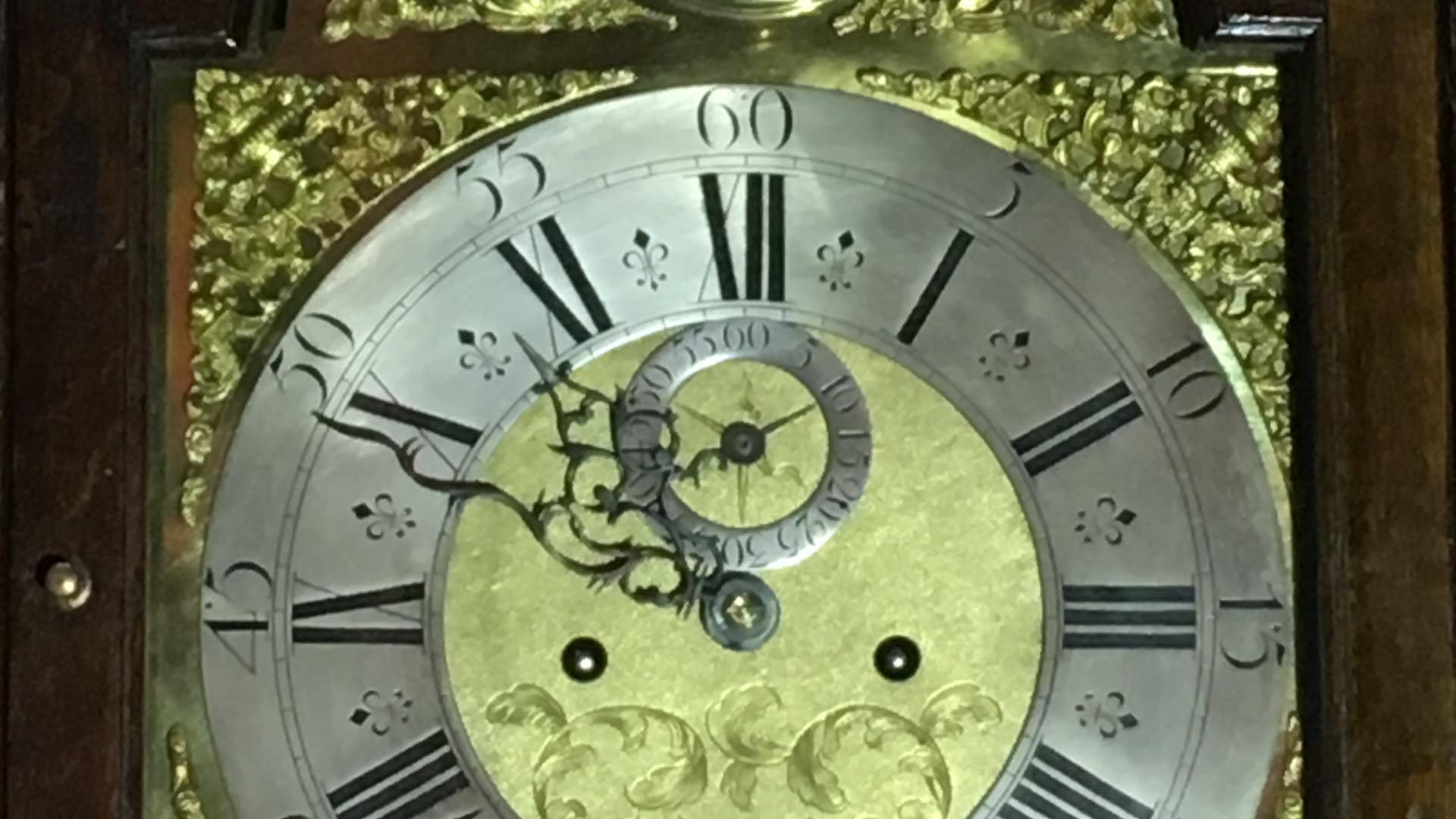 A close up of an antique clock face showing ornate hands, floral decorative motifs and Roman numerals.