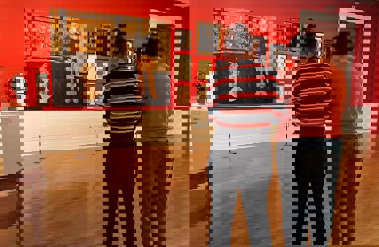 Two people stand in a red painted gallery in Graves Gallery. They are facing away from the camera, looking at a large self portrait on the wall. 