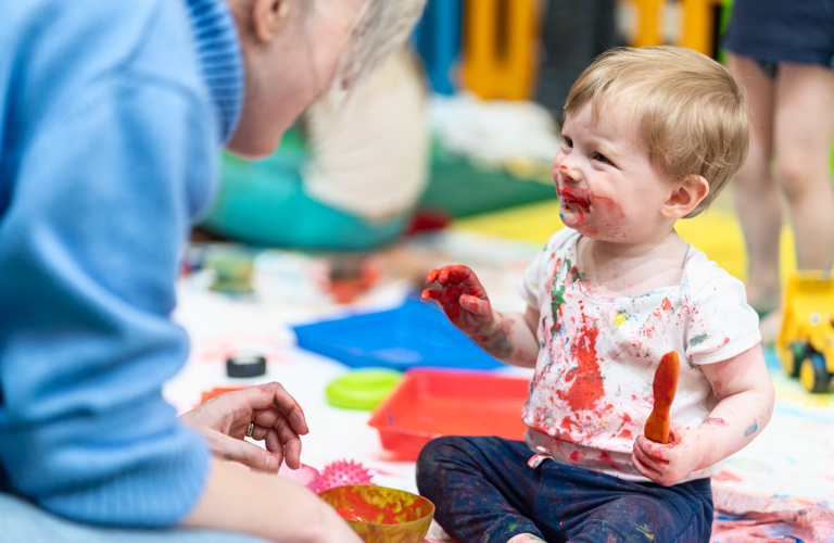 A photograph of a toddler smiling at an adult. They are holding a paintbrush and their clothes, face and hands are splattered in paint. 