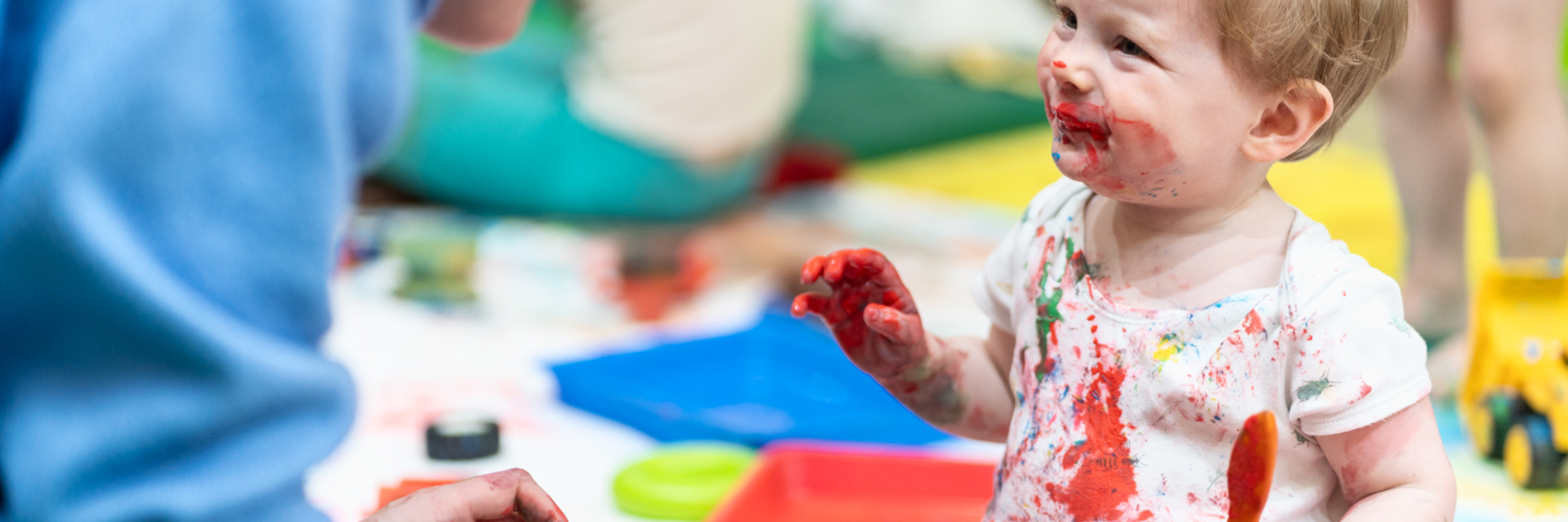 A photograph of a toddler smiling at an adult. They are holding a paintbrush and their clothes, face and hands are splattered in paint. 
