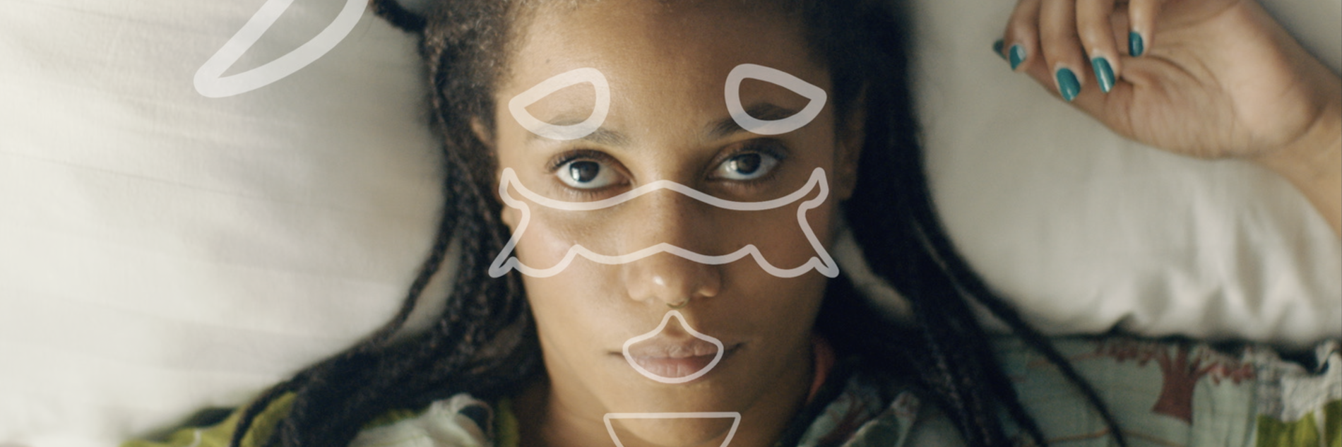 This is the alt text they want using on the image: A Black woman in her 30s lying on a bed, we see her from above and she looks up at us intensely. Her left hand with green painted nails lays next to her head on the white pillow, hair in singles on her shoulders, over a green patterned robe. She is staring not at us, but at something floating above her face, whether it is actually there or not. It is a series of symbols, formed from white outlines, in an almost-symmetrical pattern. They appear to be in motion
