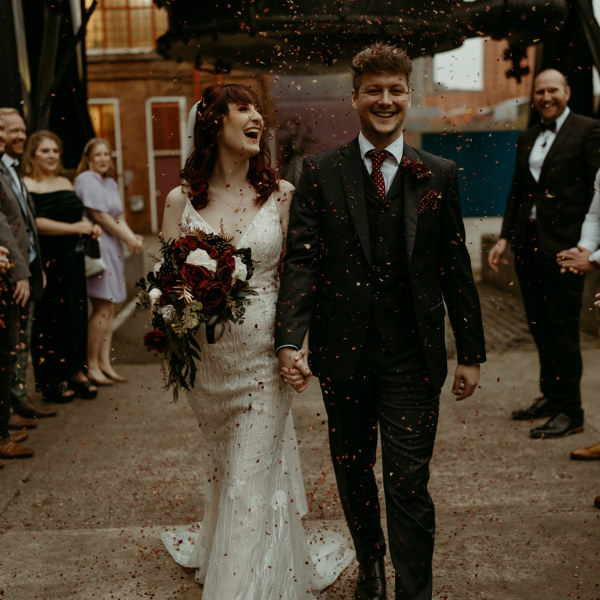 A photograph of a newlywed couple walking outside Kelham Island Museum, hand in hand, with confetti in the air. There are guests on both sides and the bride and groom are both laughing.
