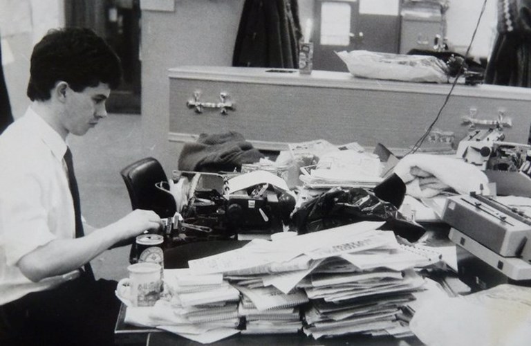 A black and white photograph showing a journalist working at a typewriter on a desk covered with papers. In the background is an empty coffin.