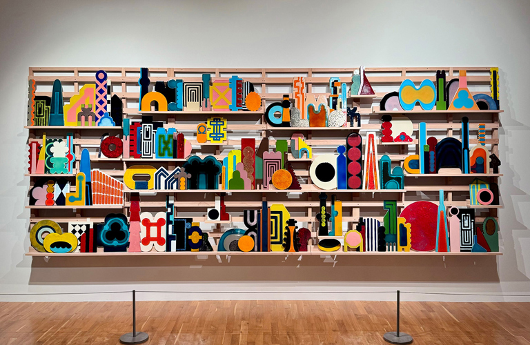 A photograph of a large, brightly coloured artwork in an art gallery. The work is made up of many small geometric shapes arranged on a wooden structure with three narrow shelves.  