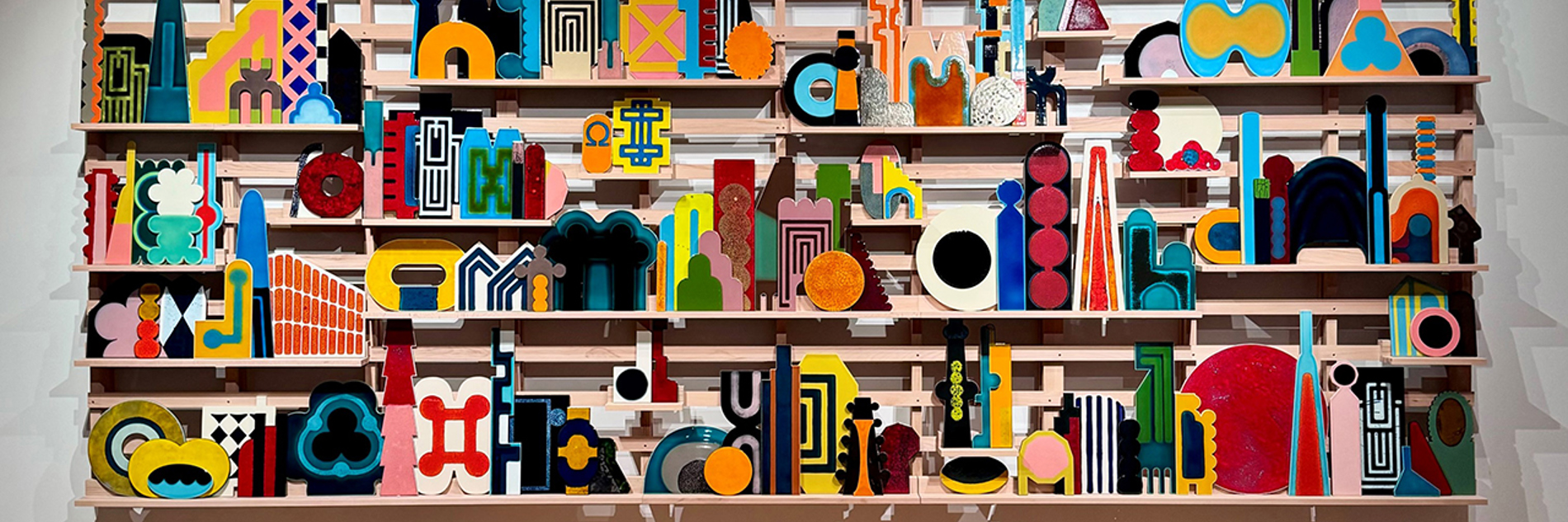 A photograph of a large, brightly coloured artwork in an art gallery. The work is made up of many small geometric shapes arranged on a wooden structure with three narrow shelves.  