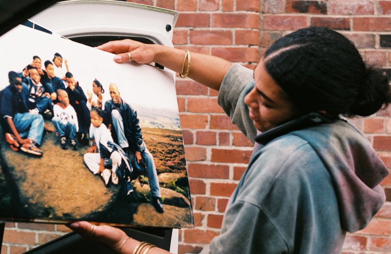 A photograph of an adult standing in front of a brick wall holding a mobile phone between their ear and shoulder and   looking at a large photograph of a group of adults and children sitting on a large stone. An outline of a car door can be seen behind the photograph. 
