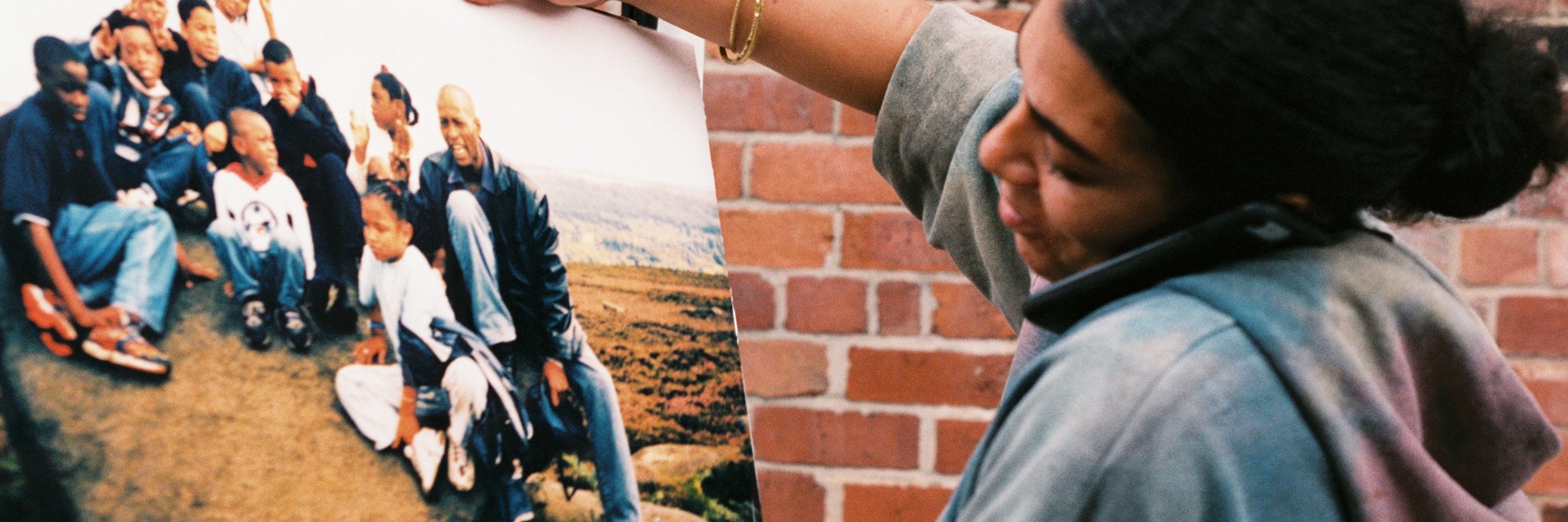 A photograph of an adult standing in front of a brick wall holding a mobile phone between their ear and shoulder and   looking at a large photograph of a group of adults and children sitting on a large stone. An outline of a car door can be seen behind the photograph. 