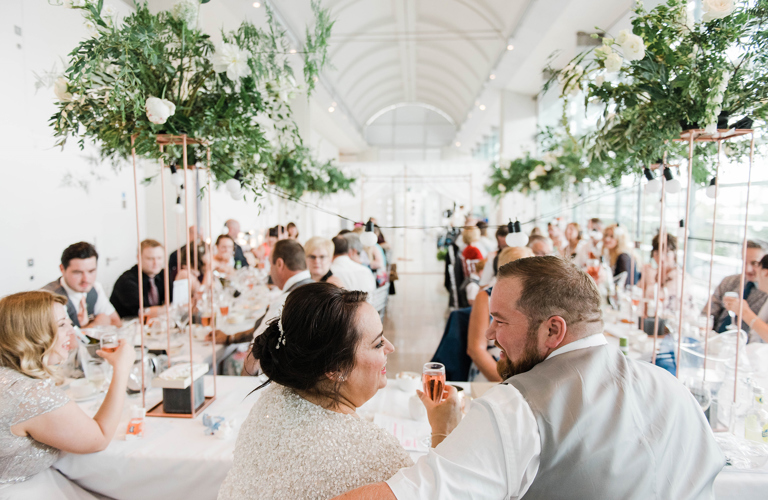 Two adults in wedding outfits sitting at a table in front of a group of formally dressed adults seated at two long tables.   
