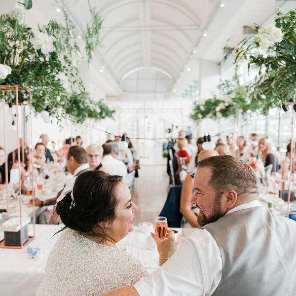 Two adults in wedding outfits sitting at a table in front of a group of formally dressed adults seated at two long tables.   