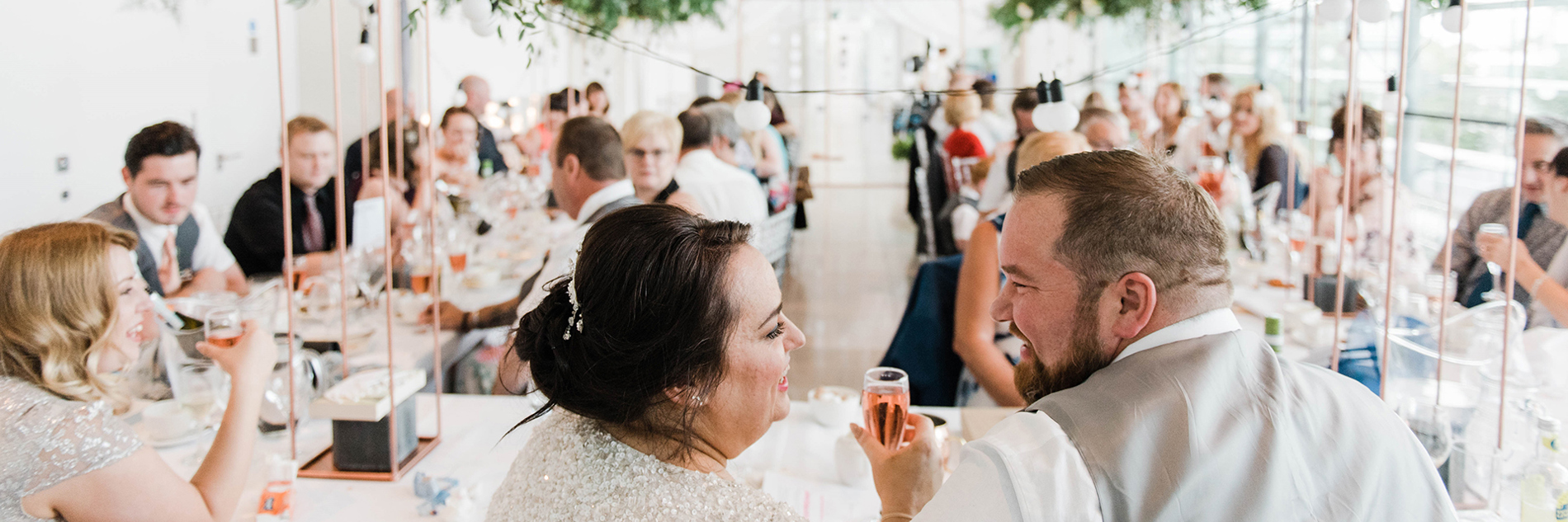 Two adults in wedding outfits sitting at a table in front of a group of formally dressed adults seated at two long tables.   