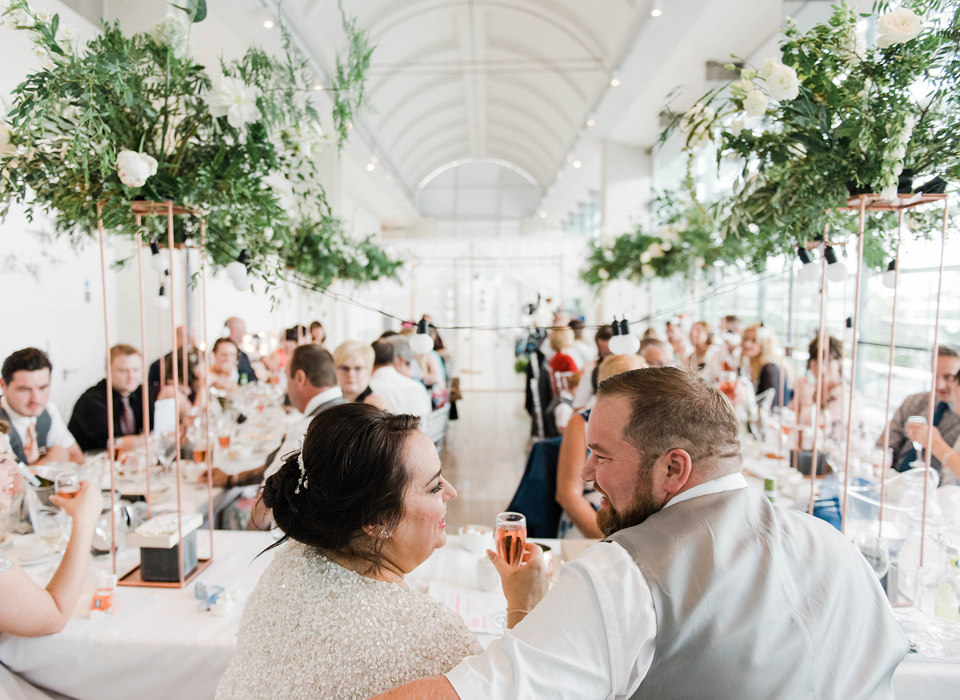 Two adults in wedding outfits sitting at a table in front of a group of formally dressed adults seated at two long tables.   