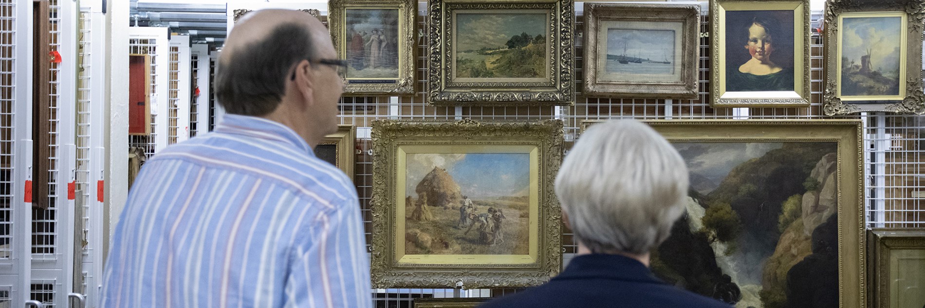 Two adults looking at a collection of framed paintings mounted on a sliding frame