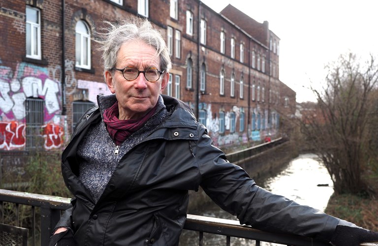 Simon Ogden stands against metal railings on a bridge over a river.  In the background is large redbrick industrial building which is covered in graffiti at ground level. On the other side of the river bank are trees.