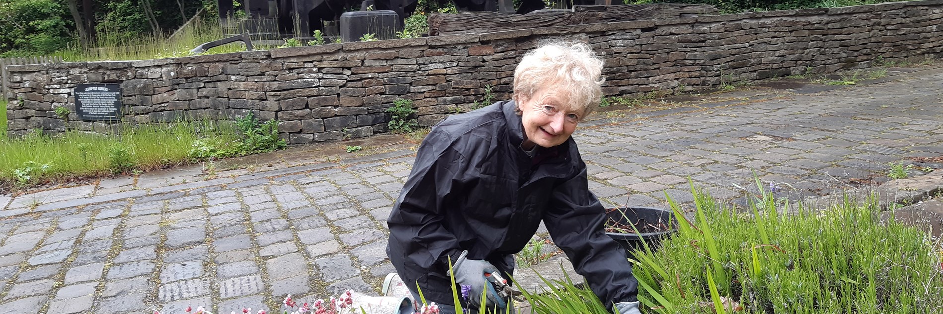 A smiling adult gardening on a cobbled walkway, in front of a cobbled wall. Some historic engineering equipment can be seen in the background 