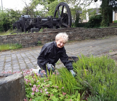 A smiling adult gardening on a cobbled walkway, in front of a cobbled wall. Some historic engineering equipment can be seen in the background