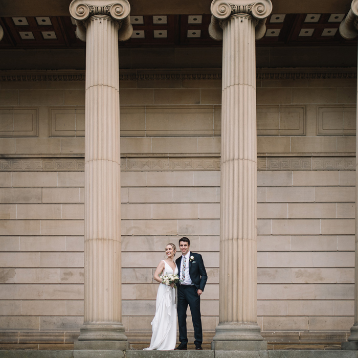 A bride in a wedding dress and a groom in a suit pose in front of a facade of a historic building with large columns