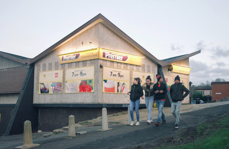 A photograph of a group of four teenagers walking down a hill walking past a Premier convenience store. They are all wearing jeans and jackets. 