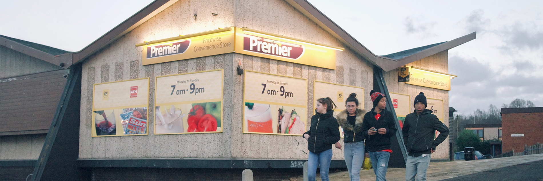 A photograph of a group of four teenagers walking down a hill walking past a Premier convenience store. They are all wearing jeans and jackets. 