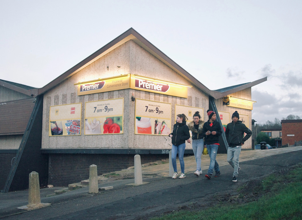 A photograph of a group of four teenagers walking down a hill walking past a Premier convenience store. They are all wearing jeans and jackets. 
