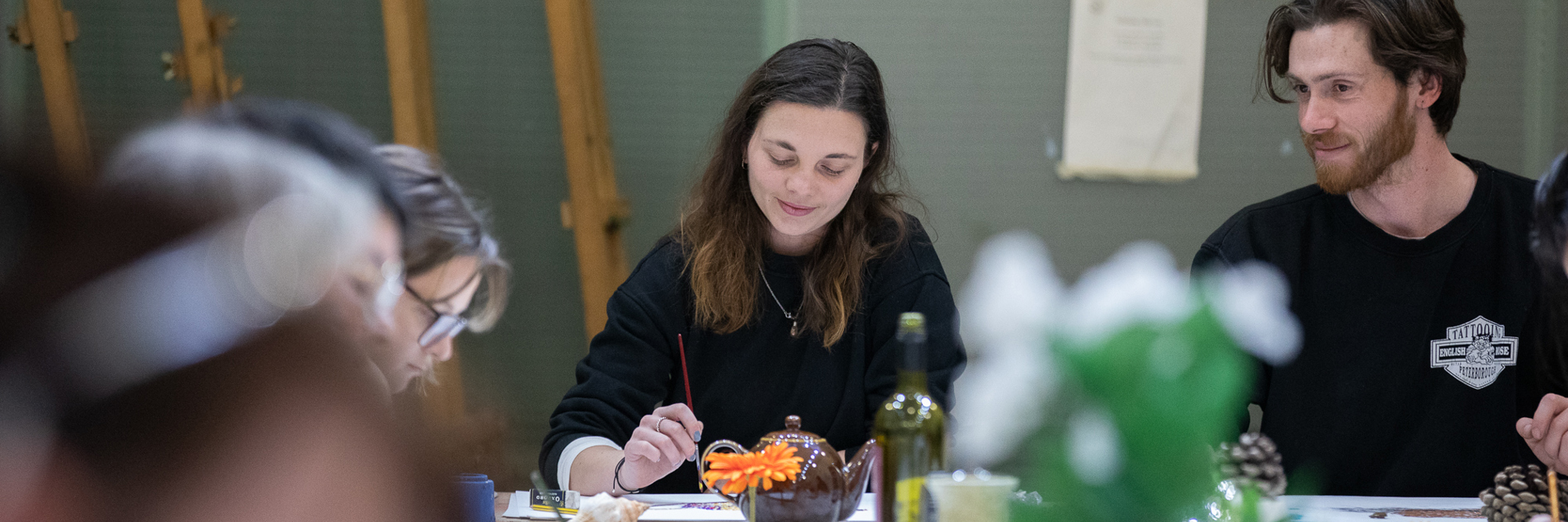 A photograph of a man and a woman sat at a table full of art materials and objects to draw. The woman is holding a paintbrush.