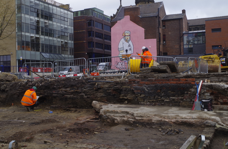 A photograph of a dig site in Sheffield City Centre. A man in an orange high vis jacket is shown in the rubble, inpsecting the sides. There are metal barriers separating the dig from the street. Another man in high vis is shown at street level. Tall office buildings are visible in the background, and a large mural of a grandma is shown on a pink background on one of the buildings.