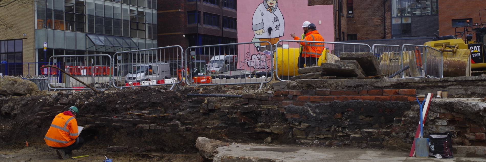 A photograph of a dig site in Sheffield City Centre. A man in an orange high vis jacket is shown in the rubble, inpsecting the sides. There are metal barriers separating the dig from the street. Another man in high vis is shown at street level. Tall office buildings are visible in the background, and a large mural of a grandma is shown on a pink background on one of the buildings.