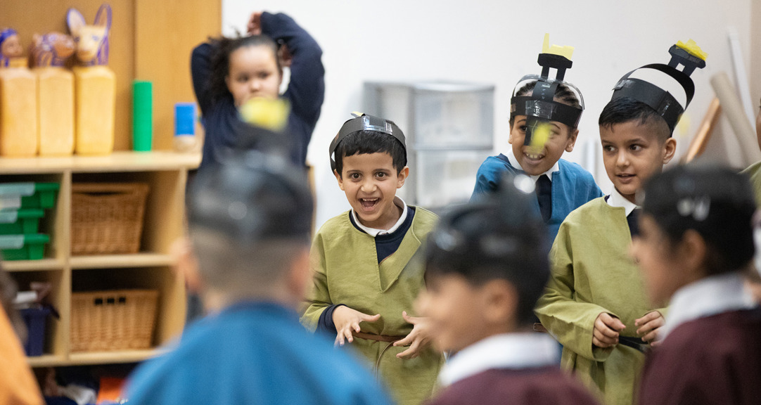Seven smiling primary school aged children taking part in a school session. They are dressed as Anglo Saxons and wearing brightly coloured tunics and paper helmets which they have made in the session.