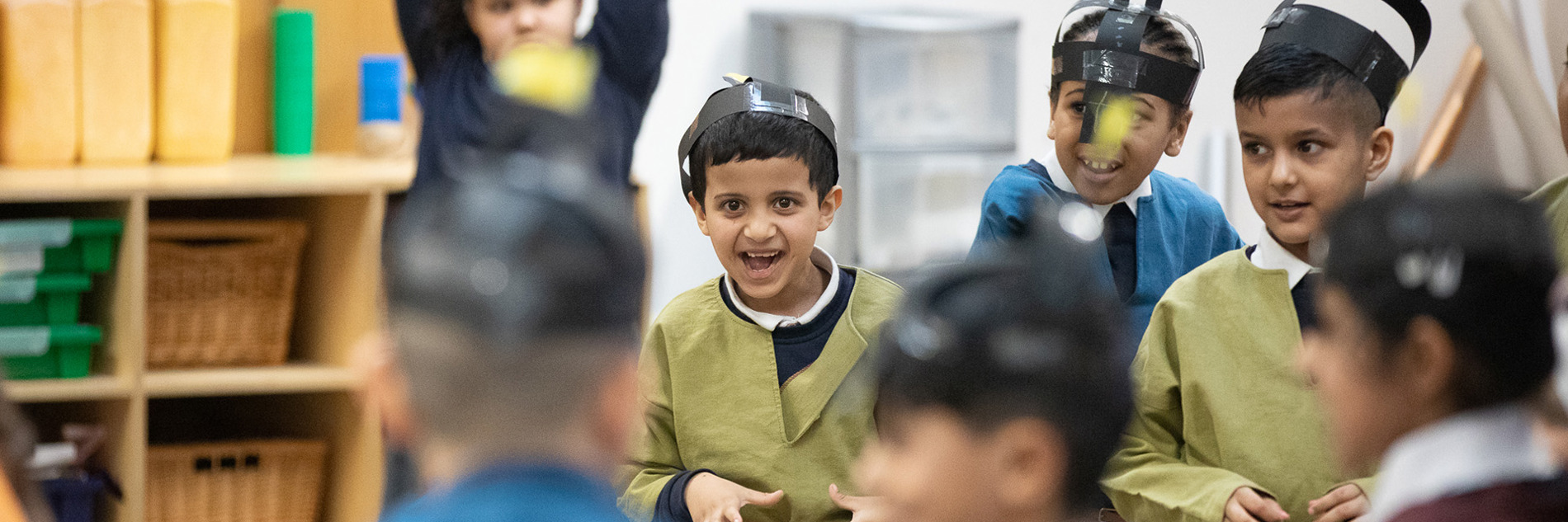 Seven smiling primary school aged children taking part in a school session. They are dressed as Anglo Saxons and wearing brightly coloured tunics and paper helmets which they have made in the session.
