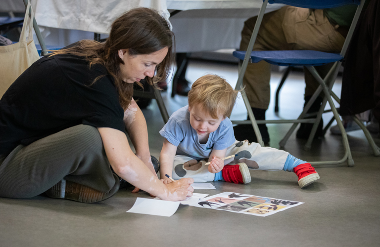 A photograph of a woman and a young boy sat on the floor with a sheet of bird images in front of them. They are both sketching on another piece of paper.
