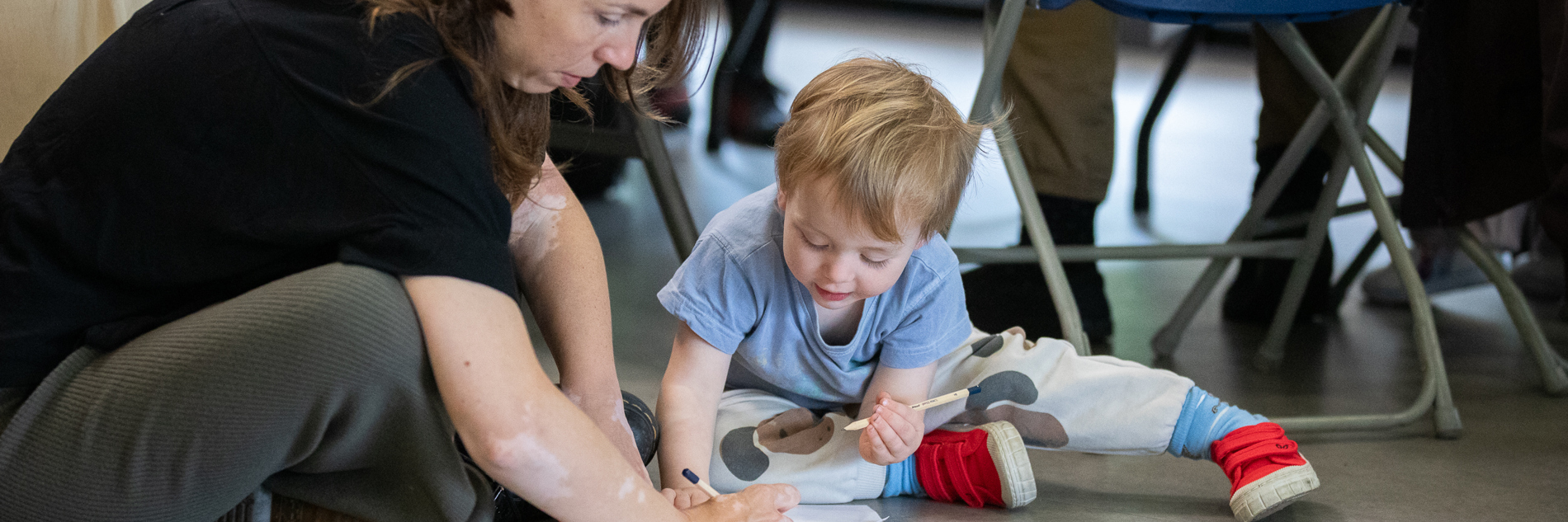 A photograph of a woman and a young boy sat on the floor with a sheet of bird images in front of them. They are both sketching on another piece of paper.