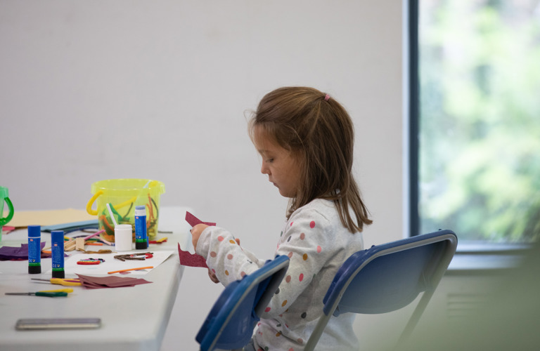 A photograph of a young girl sat at a table of crafting materials, carefully cutting a circle out of a sheet of red paper