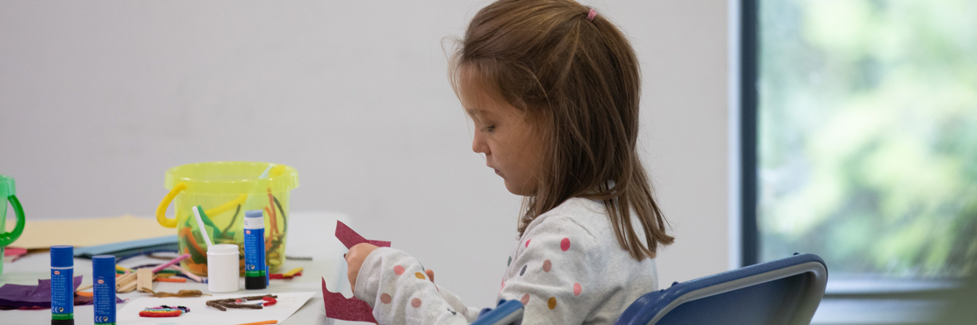 A photograph of a young girl sat at a table of crafting materials, carefully cutting a circle out of a sheet of red paper