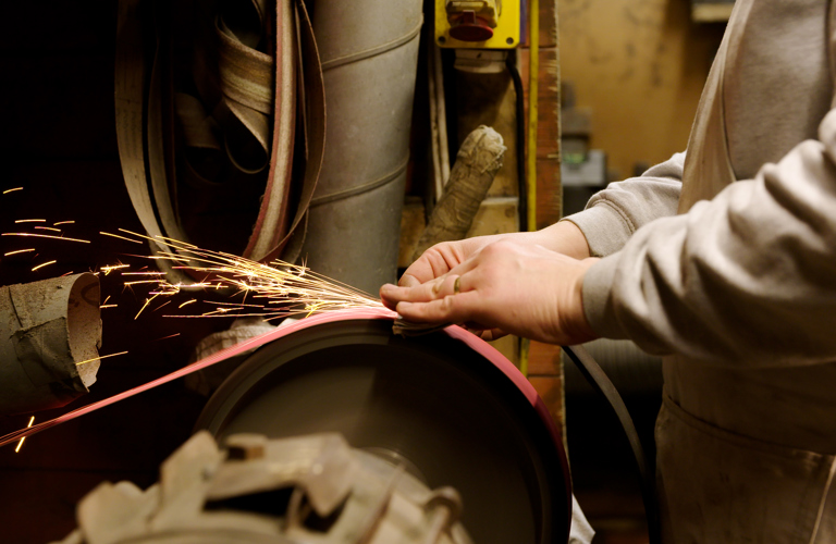 A torso and hands of a person using a grinding wheel and creating sparks. In the background there is a workshop with tubing, switches and machinery. 