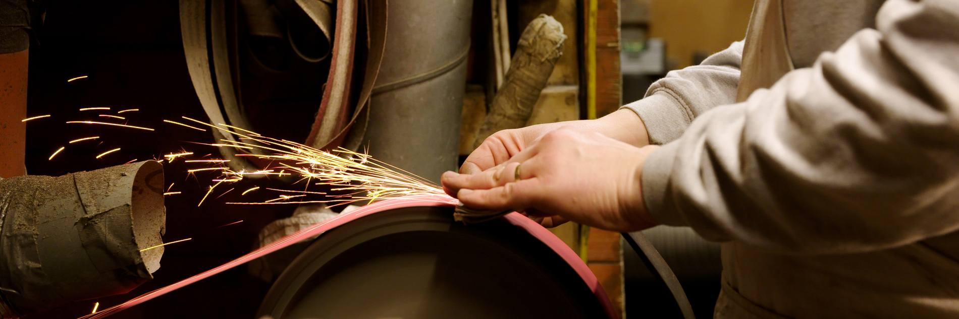 A torso and hands of a person using a grinding wheel and creating sparks. In the background there is a workshop with tubing, switches and machinery. 